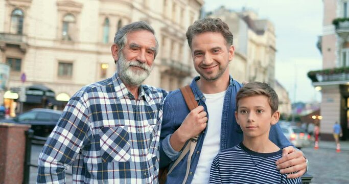 Portrait of happy cheerful Caucasian handsome father, old grandfather and teen son grandson smiling to camera at street in town. Family o two joyful men and boy. Different ages and generations.