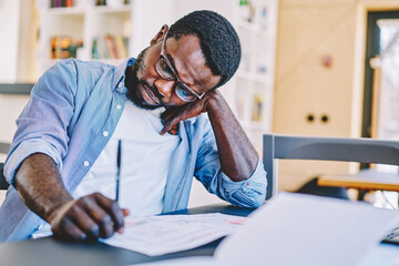 Serious african american male student puzzled on doing homework task sitting at desktop, concentrated serious hipster guy planning costs writing in notepad and pondering on idea for solution.