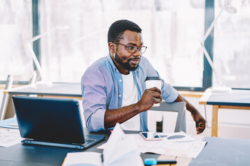 Serious african american guy holding cardboard cup thinking about job on work break, pensive dark skinned male freelancer sitting at desktop with coffee thoughtfully looking away in coworking space.