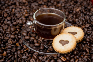 a cup of black coffee with a dish and two cookies which have a chocolate heart in the middle of them, black coffee beans in the background