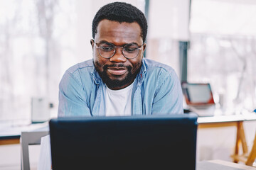 Clever african american male in eyewear for vision protection watching webinar for education on laptop computer,serious dark skinned hipster guy reading information from web page making banking .