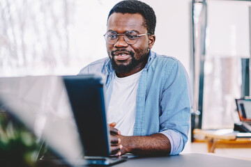 Smart african american dark skinned man watching tutorial on laptop computer wear eyeglasses for vision protection, dark skinned millennial guy satisfied with wifi connection enjoying movie online. - obrazy, fototapety, plakaty