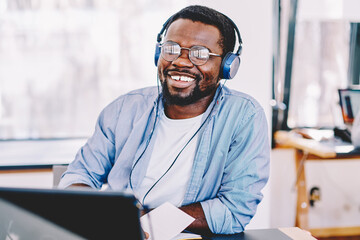 Cheerful african american hipster guy enjoying music in modern headphones and online radio broadcasting on laptop computer, dark skinned male student entertaining with litening playlist on netbook.