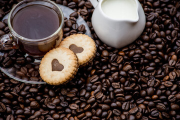  a cup of black coffee, two cookies and milk beside it. black coffee beans in the background