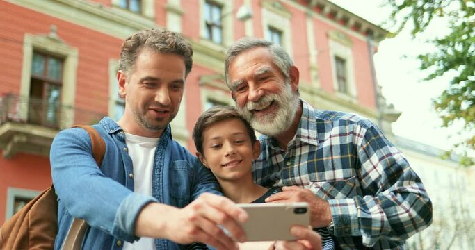 Cute teen Caucasian boy with father and senior grandfather taking selfie photo at street with smartphone. Old man with beard with adult son and small grandson posing while making photos on phone.