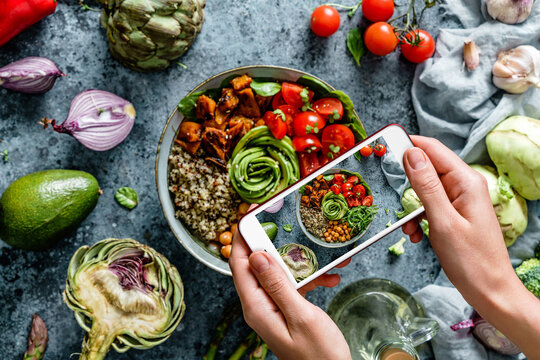 Girl's hands taking photo of Buddha bowl salad with chickpeas, quinoa, tomatoes, arugula, avocado, sprouts by smartphone. Healthy vegan food, clean eating, dieting, top view