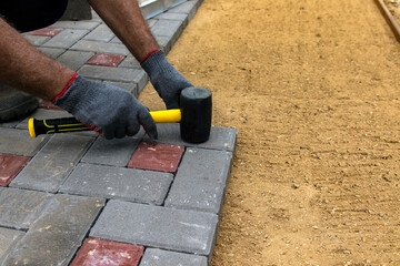 A craftsman in gloves with a rubber mallet lays concrete blocks on a gravelly sand base. Laying concrete blocks on the sidewalk.