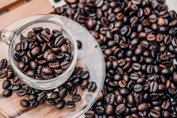 coffee cup and saucer  filled with black coffee beans, both placed on a wooden platform with a heap of coffee beans beside it. white background