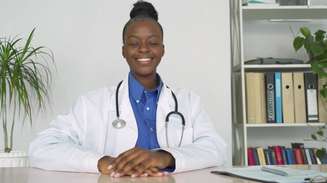 Confident smiling african female doctor looking at camera sit at workplace. Afro american woman professional therapist wear white lab coat and stethoscope posing at work desk. Black physician portrait