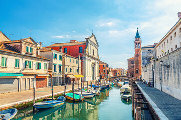 Chioggia cityscape with narrow water canal Vena with moored multicolored boats, Chiesa dei Filippini church, tower of Parrocchia di San Giacomo Apostolo building and brick bridge, Northern Italy