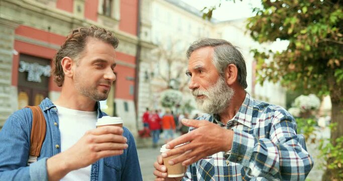 Caucasian handsome young adult man talking with old father while walking in city street and drinking coffee to-go. Two me of different ages and generations having nice conversation outside in town.