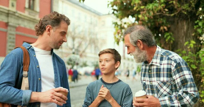 Handsome Caucasian adult man with old father drinking coffee to-go at street and talking with little teen son. Boy talk with dad and grandfather while walking outdoors in town.