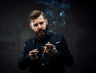 Elegantly dressed bearded male with tattoos on his neck and hand lights a cigar. Brutal young...