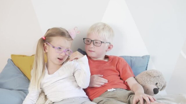 Funny kids with glasses. Brother and his little sister with glasses sit on the bed.
