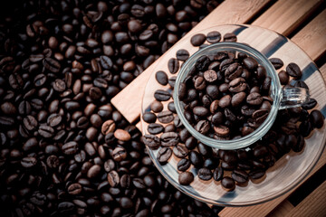 glass coffee cup and a saucer both filled with coffee beans, with black coffee beans in the background