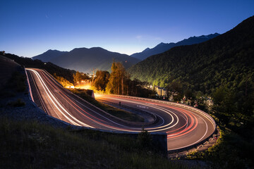 A winding road in the mountains. A trail from the car headlights. Mountains. Evening lighting. - obrazy, fototapety, plakaty