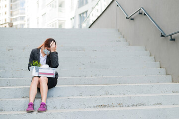 A woman with a box of personal belongings sits on the stairs outside and folded her hands in despair on head. Female employee in medical mask fired due to financial crisis in coronovirus. Unemployment