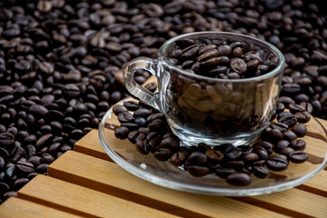 glass coffee cup and a saucer both filled with coffee beans, with black coffee beans in the background