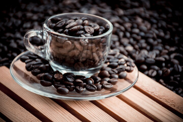 glass coffee cup and a saucer both filled with coffee beans, with black coffee beans in the background