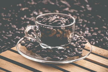glass coffee cup and a saucer both filled with coffee beans, with black coffee beans in the...