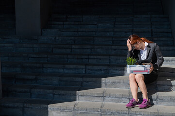 Fototapeta premium Depressed woman dressed in medical mask is fired and is sitting on the stairs with a box of personal belongings. Female office worker in suit and sneakers outdoors. Unemployment in the economic crisis
