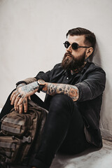 Travel and education concept. Bearded hipster guy in sunglasses with backpack sitting on a floor in studio
