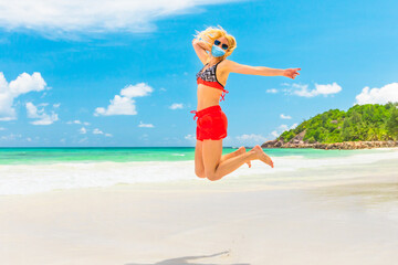 Woman in bikini jumping on seashore of Anse Kerlan Seychelles, with surgical mask during Covid-19. Praslin, Seychelles freedom in tropical sea in coronavirus quarantine. Holidays in pandemic.