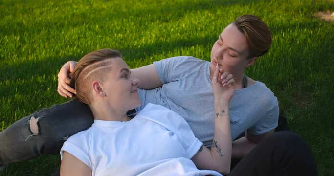 Close up portrait of female same-sex couple lying on the grass. Two pretty stylish girls with short creative hairstyles hug and kiss each other in love.  4k slow motion 50 fps