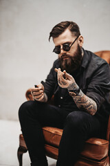 Cool tattooed bearded man lights a cigar while sitting on a vintage chair in studio