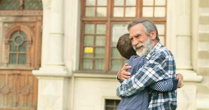 Caucasian happy senior grandfather meeting and hugging his cute joyful teen grandson at street in city. Cheerful old gray-haired man and teenage boy meet outdoor.