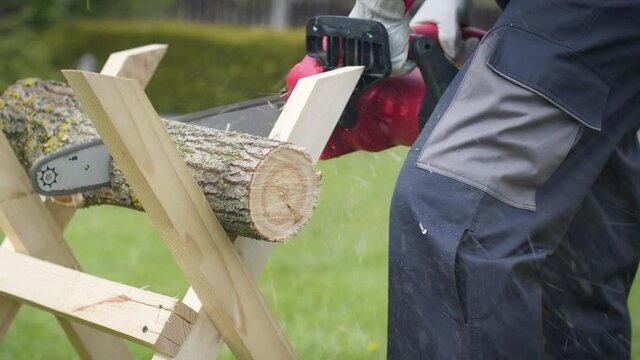 Caucasian man in gloves saws firewood on sawhorses with an electric saw at his home site. Harvesting firewood for the winter. Men's work. Wooden logs. Close-up.