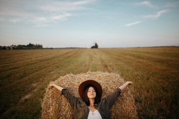 Stylish girl relaxing on hay bale in summer field in sunset. Portrait of young sensual woman in...