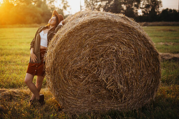 Stylish girl relaxing on hay bale in summer field in sunset. Portrait of young sensual woman in resting at haystack, atmospheric tranquil moment. Countryside slow life