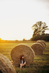 Stylish girl relaxing at hay bale in summer field in sunset. Young woman in hat sitting at...
