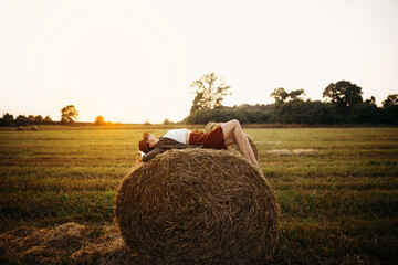 Stylish girl relaxing on hay bale in summer field in sunset. Young woman in hat resting on hay in...