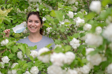 Happy red-haired woman takes off a medical mask while standing next to a blossoming apple tree in a...