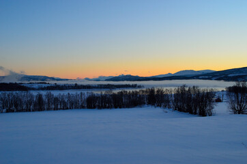 ice fog over calm fjord with strong colorful sunset sky and snowy mountain range in winter