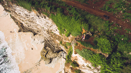 Top view aerial photo from flying drone of a young loving couple are enjoying amazing landscape of Indian Ocean, while are standing on a green cliff during their beautiful honeymoon in Bali Indonesia