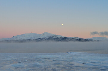 pink sunset sky and full moon over winter fjord with dense ice fog and reflection on water surface