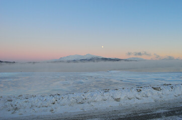 pink sunset sky and full moon over winter fjord with dense ice fog and reflection on water surface
