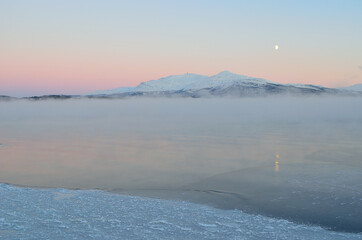 pink sunset sky and full moon over winter fjord with dense ice fog and reflection on water surface