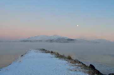 pink sunset sky and full moon over winter fjord with dense ice fog and reflection on water surface