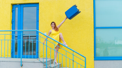 A happy red-haired woman in a yellow dress is walking down the stairs and joyfully swing a blue suitcase. The girl is preparing for the journey. Summer vacation concept.