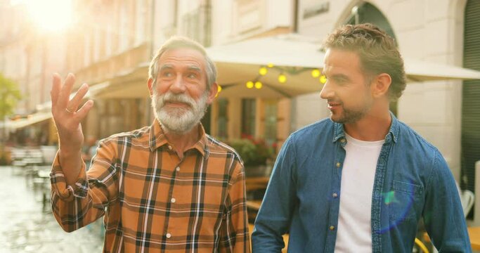 Two male generations walking the street in town and talking. Father and son strolling in city and talk with smiles outdoor. Senior dad sharing his wisdom with young man outdoor. Men of different ages.