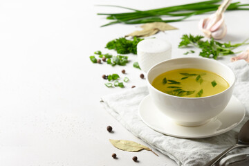 Chicken broth with spices, greens, garlec, parsley, onion in white bowl on white table. Copy space. Close up.