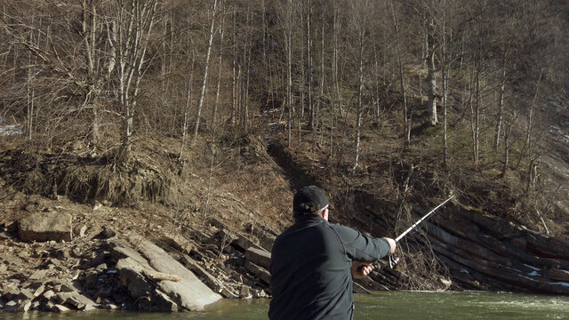 Adult man fishing at mountain stream. Throws a fishing rod into the river at the rocky terrain. Rolling the reel with fishing line.