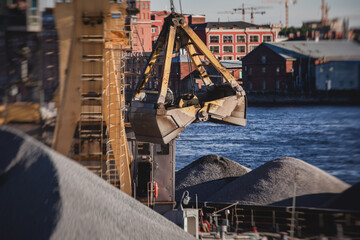 Naklejka premium Bulk-handling crane unloading sand, road metal and gravel from cargo vessel ship, heavy vehicle and portal crane loader working with bulk materials in dock terminal
