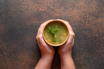 Chicken broth with greens in craft paper containers in child hand on brown background. Soup to go,...