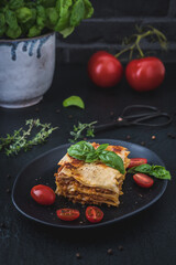 Piece of homemade vegetarian soy lasagne on a black plate, tomatoes, herbes and basil on dark background, vertical