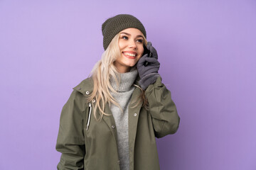 Teenager blonde girl with winter hat over isolated purple background keeping a conversation with the mobile phone